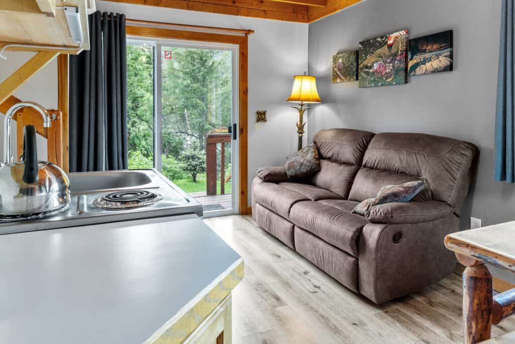 Cozy living room inside Kenai River Drifters Lodge with a brown sofa, lamp, and a view of lush greenery through a sliding door.