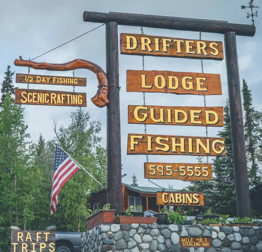 Wooden sign for Kenai River Drifters Lodge with fishing, rafting, and lodge info. Nature background, American flag.