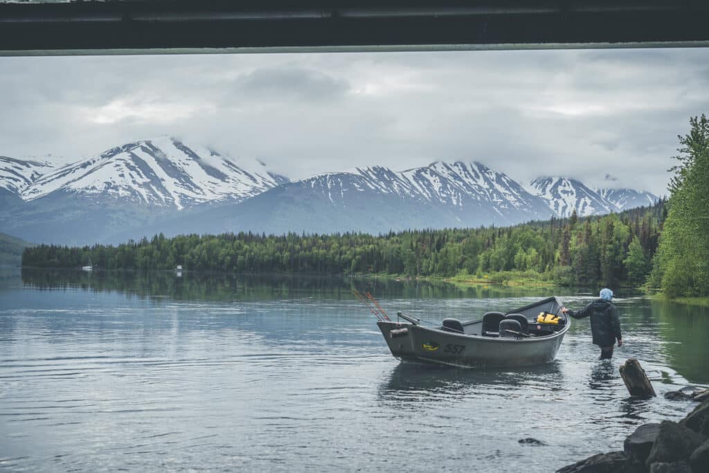 Serene lake scene with snow-capped mountains and a fisherman launching a boat in lush green surroundings.