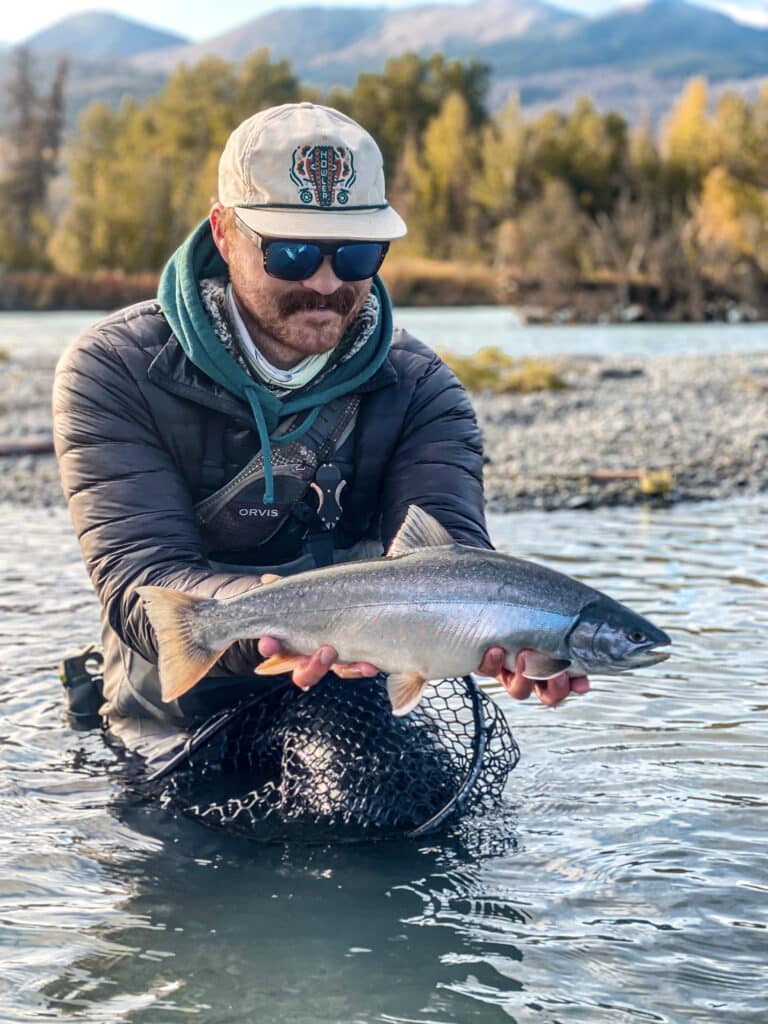 A man holding a large fish in a river with mountains and trees in the background.