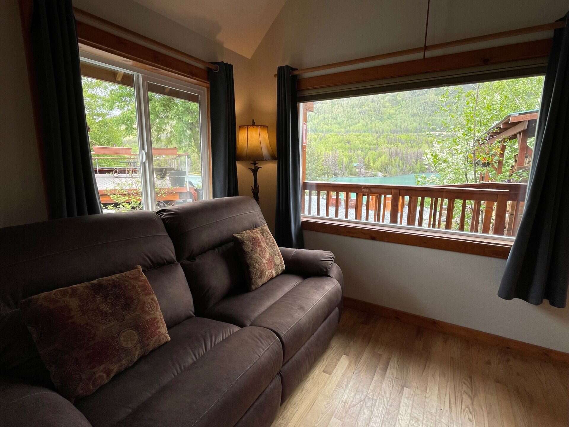 Cozy living room with large windows overlooking the Kenai River and surrounding forest, featuring a comfortable dark brown couch and warm lighting.