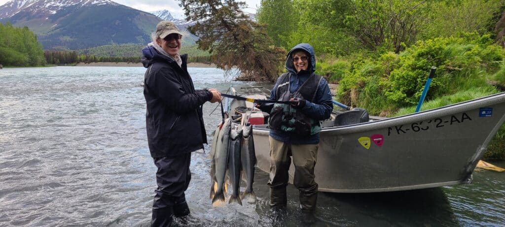 Two anglers standing on the riverbank with a boat in the background, proudly holding a stringer of freshly caught salmon from the Kenai River
