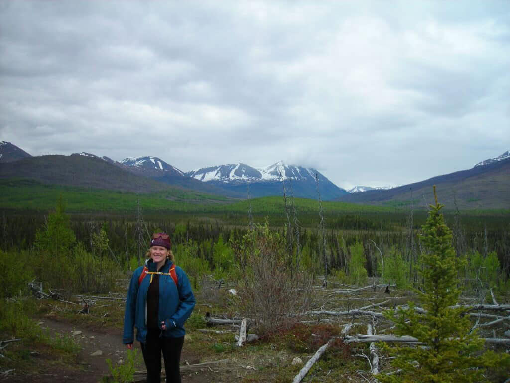 Serene Alaskan mountain landscape with a smiling woman hiking in the foreground. Snow-capped peaks and lush green forest surround her.