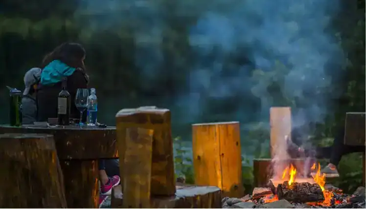 Lodge outdoor fire pit with a woman sitting on a wood bench in Kenai River Drifters Lodge, Alaska, enjoying nature.