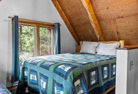 Cozy mountain cabin bedroom with wood-paneled ceiling and large window overlooking forest.