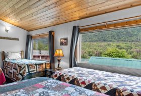 Cozy upstairs bedroom with wooden accents and large windows overlooking lush green forest.
