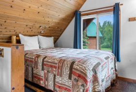Cozy bedroom with wooden ceiling, large window with blue curtains, and a quilted bedspread at Kenai River Drifters Lodge.