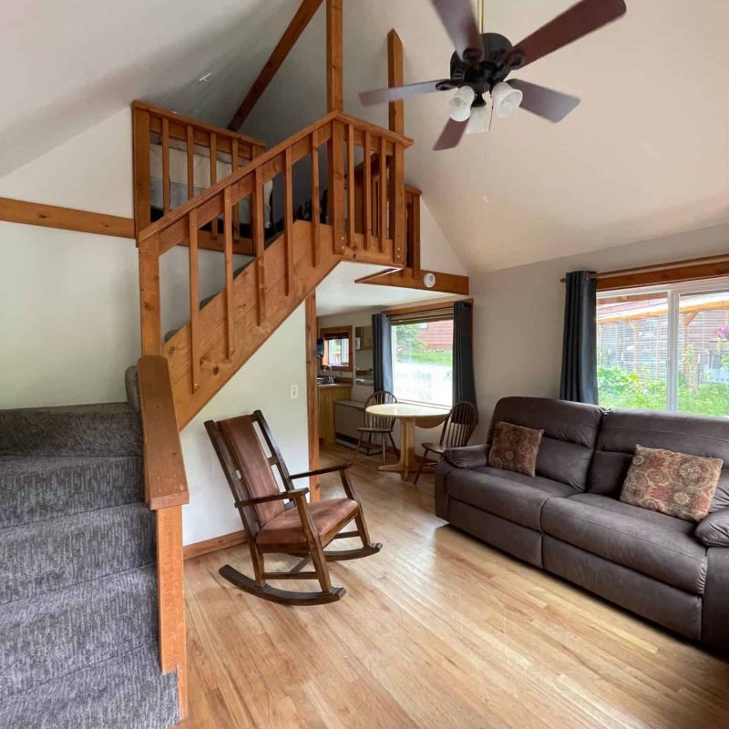 Cozy living room with wood accents and a loft area, featuring a rocking chair, sofa, and natural light from large windows.
