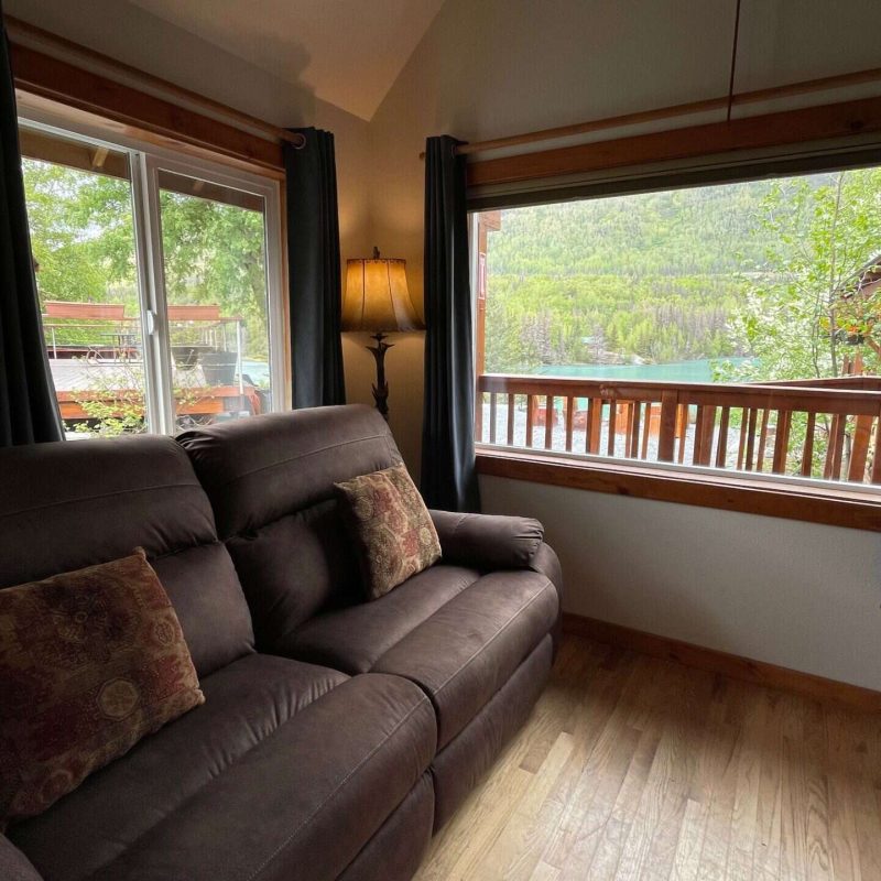 Cozy living room with large windows overlooking the Kenai River and surrounding forest, featuring a comfortable dark brown couch and warm lighting.