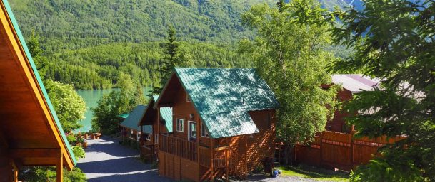 Cozy wooden cabins with green metal roofs nestled in lush trees near the Kenai River and surrounded by snow-capped mountains.