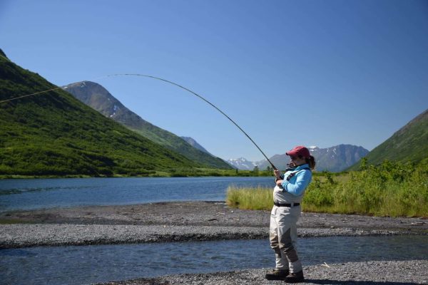 Fishing woman casting in Kenai River surrounded by mountains, lush greenery, and clear blue sky.