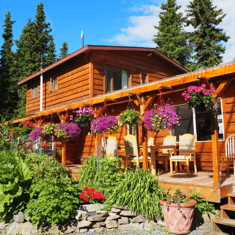 Colorful wooden lodge with hanging flower baskets and outdoor seating amid lush greenery at Kenai River Drifters Lodge.